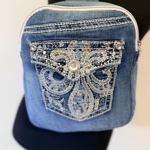 May include: A denim crossbody bag with a decorative pocket. The pocket features intricate silver embroidery and is embellished with clear and gold-colored rhinestones. The bag has a white zipper and strap.