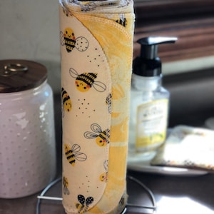May include: A roll of reusable paper towels with a bee pattern, displayed on a metal stand. The towels feature a cream background with black and yellow bees. The edge of the roll reveals a yellow floral pattern. A soap dispenser and jar are in the background.