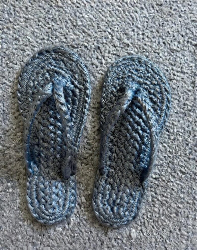 May include: A pair of slate blue flip-flops crafted from thick, braided cord material. The footwear features a rounded toe bed and a Y-shaped strap design, both constructed with a consistent, textured crochet-like weave pattern throughout.