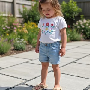 May include: A white short-sleeved t-shirt featuring a colorful embroidered floral pattern on the chest, paired with light blue denim shorts with a cuffed hem and beige woven flip-flops with a textured braided strap design.