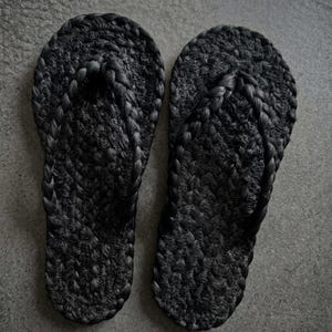 May include: A pair of black flip-flops featuring a textured, braided rope construction. The footwear has a rounded toe shape and a thick, woven strap design that connects to the sole, creating a uniform, dark appearance throughout the entire item.