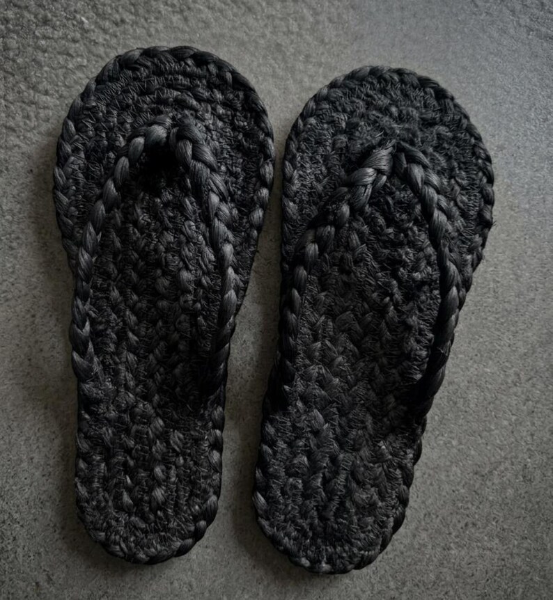 May include: A pair of black flip-flops featuring a textured, braided rope construction. The footwear has a rounded toe shape and a thick, woven strap design that connects to the sole, creating a uniform, dark appearance throughout the entire item.