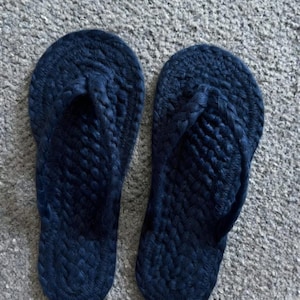 May include: A pair of dark navy blue flip-flops featuring a textured, braided rope construction. The footwear has a rounded toe shape and a thick, woven strap design that secures the foot, showcasing a consistent dark blue color throughout the entire surface.