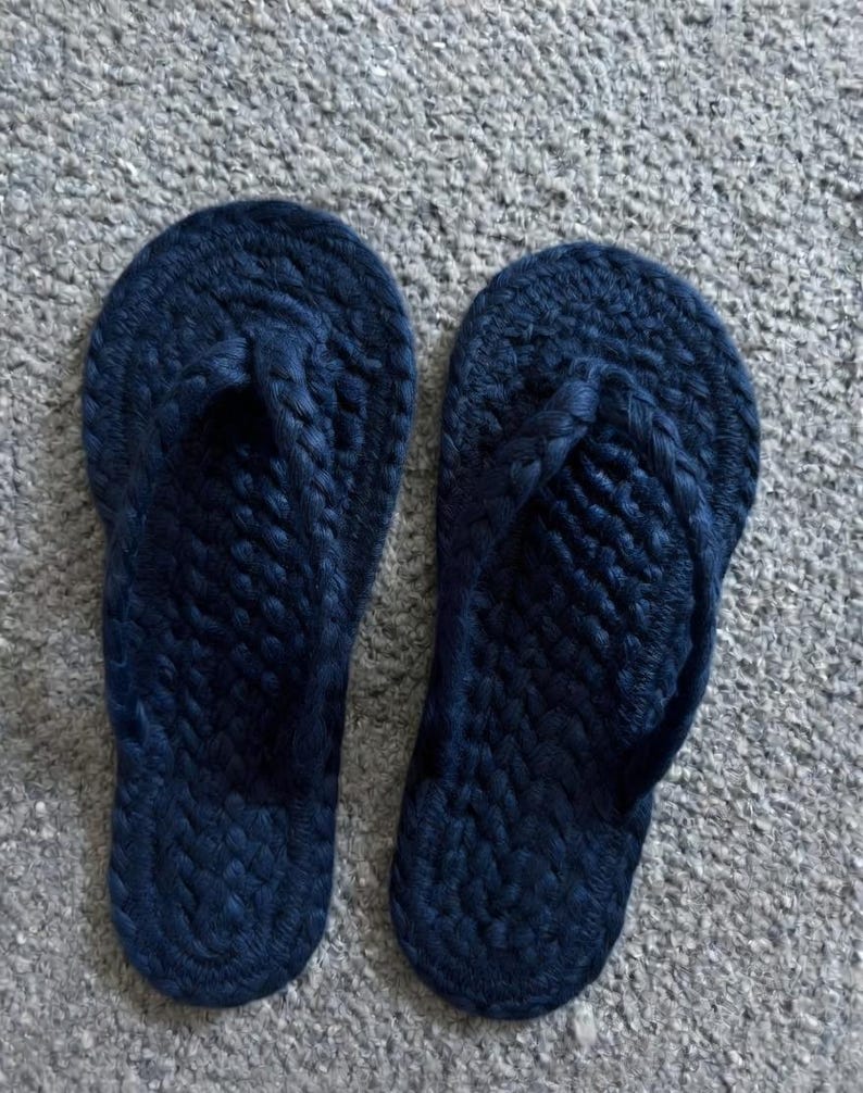 May include: A pair of dark navy blue flip-flops featuring a textured, braided rope construction. The footwear has a rounded toe shape and a thick, woven strap design that secures the foot, showcasing a consistent dark blue color throughout the entire surface.