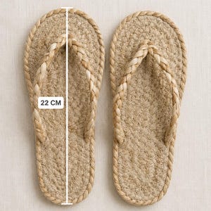 May include: A pair of tan flip-flops crafted from braided natural fiber rope. The sandals feature a woven sole and a braided strap design. A white measurement line is overlaid on the left sandal, clearly indicating a length of 22 cm.