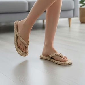 May include: A pair of beige flip-flops featuring a braided strap design and a textured, woven-style footbed. The sandals have a simple thong construction with a flat sole, showcasing a neutral tan color throughout the entire footwear design.