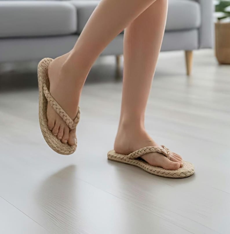 May include: A pair of beige flip-flops featuring a braided strap design and a textured, woven-style footbed. The sandals have a simple thong construction with a flat sole, showcasing a neutral tan color throughout the entire footwear design.