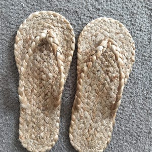 May include: A pair of flip-flops crafted from natural tan woven jute fibers. The footwear features a braided strap design and a textured, braided sole construction, showcasing a consistent light brown color throughout the entire material.