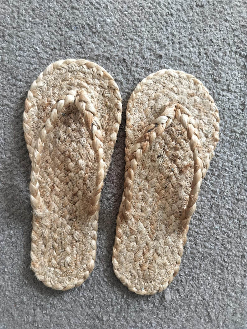 May include: A pair of flip-flops crafted from natural tan woven jute fibers. The footwear features a braided strap design and a textured, braided sole construction, showcasing a consistent light brown color throughout the entire material.