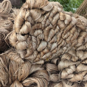 May include: A large, dense pile of raw, light brown jute fibers. The material consists of long, coarse, and fibrous strands bundled together in overlapping, curved, and tangled loops, creating a highly textured, organic, and fibrous surface.