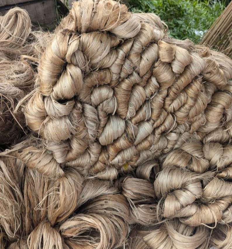 May include: A large, dense pile of raw, light brown jute fibers. The material consists of long, coarse, and fibrous strands bundled together in overlapping, curved, and tangled loops, creating a highly textured, organic, and fibrous surface.