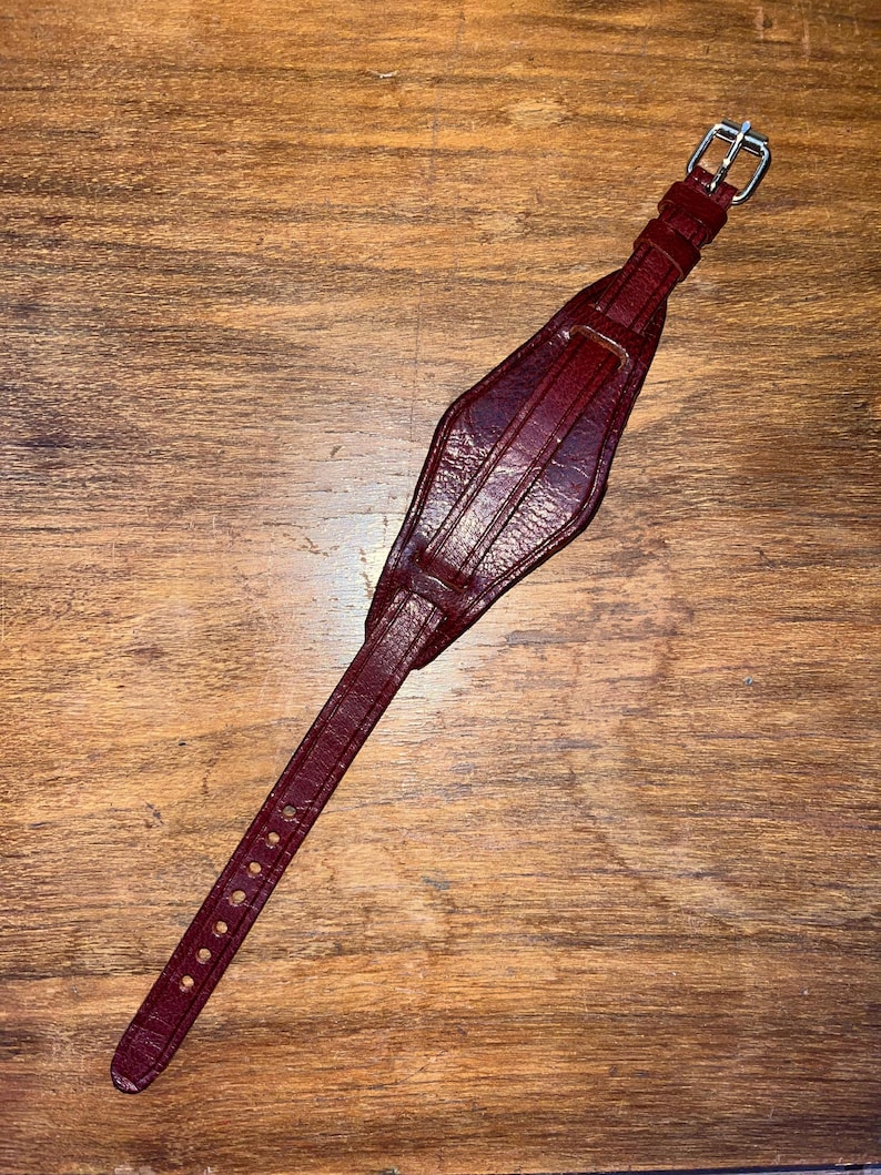 May include: A dark red leather watch strap with a silver buckle. The strap is designed with a unique, diamond-shaped section in the middle.