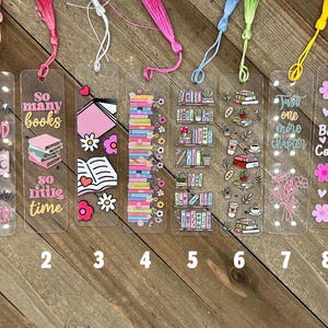 May include: Eight clear acrylic bookmarks with colorful tassels. Each bookmark features unique designs, including stacks of books, floral accents, and inspirational quotes like "So many books, so little time." The bookmarks are numbered 1 through 8.