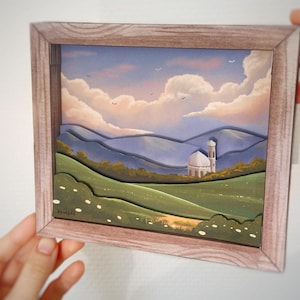 Könnte beinhalten: Gerahmtes Landschaftsbild mit einer ruhigen Szene mit grünen Hügeln, einem weißen Kuppelgebäude und einem weichen, bewölkten Himmel. Der Rahmen besteht aus hellbraunem Holz. Das Kunstwerk wird von Händen gehalten.
