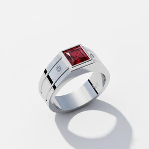 Puede incluir: Un anillo plateado con una gema roja cuadrada en el centro. El anillo tiene un diseño moderno con dos pequeñas piedras redondas y un acabado pulido. El anillo está sobre un fondo blanco.