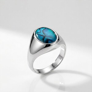 Könnte beinhalten: Ein polierter Silberring mit einem ovalen, irisierenden blau-grünen Stein. Der Ring hat einen glatten, abgerundeten Ring und eine Lünettenfassung. Der Ring ist innen mit "925 JFM" markiert.