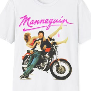 Może przedstawiać: Biała koszulka z grafiką z filmu "Mannequin". Grafika przedstawia parę na czerwonym motocyklu. Słowo "Mannequin" jest napisane różowym pismem u góry obrazu.