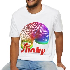 May include: White t-shirt with a vibrant rainbow Slinky graphic. The Slinky is shown in a coiled spring form, with the word "Slinky" in red text. The t-shirt has a standard crew neck.