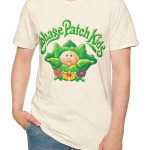 Op de afbeelding: Crèmekleurig T-shirt met de tekst "Cabbage Patch Kids" in groen, met een afbeelding van een baby in een koolblad omringd door bloemen. Het shirt heeft een gespikkelde textuur en een ronde hals.