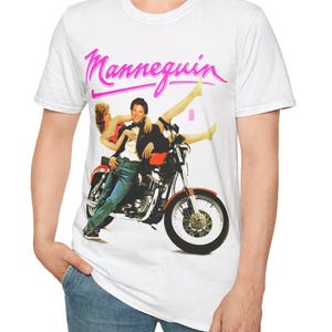 Może przedstawiać: Biała koszulka z grafiką pary na motocyklu i napisem "Mannequin" w różowym skrypcie. Projekt ma styl retro i vintage.