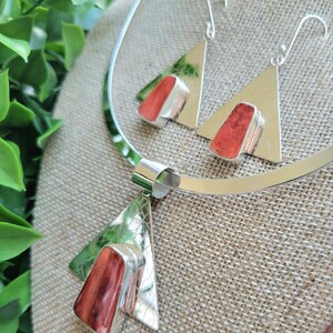 May include: A silver necklace and earrings set with triangular pendants featuring red coral stones. The necklace has a simple, wide band and the earrings have a hook closure.