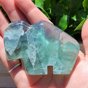 May include: A green fluorite crystal carved into the shape of a bison. The crystal is translucent and has a light green color with white veins running through it.