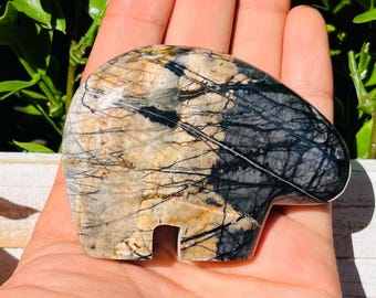 May include: A carved stone bear figurine with a unique pattern of black, tan, and gray veining. The bear has a simple, stylized design with a cutout for the legs. The stone is polished and smooth, with a natural, earthy aesthetic.