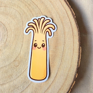May include: A yellow sticker of a cartoon mushroom with a smiling face and pink cheeks. The mushroom has a long stem and a crown of wavy lines. The sticker is outlined in white and is on a wooden surface.