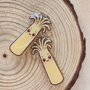Peut inclure: Deux épingles en forme de champignon avec des visages souriants. Les épingles ont un corps jaune et un dessus brun et ondulé. Elles sont posées sur un fond en bois avec un motif circulaire.