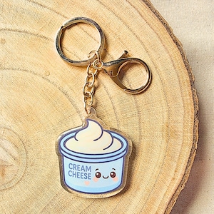 May include: A clear acrylic keychain featuring a cartoon cream cheese container with a smiling face. The container is light blue with the words "CREAM CHEESE" and a swirl of cream cheese on top. The keychain has a gold-colored ring, chain, and clasp.
