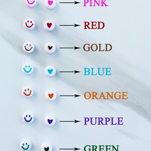 Op de afbeelding: Een set witte kralen met smiley-gezichten en harten in verschillende kleuren. De kleuren zijn roze, rood, goud, blauw, orange, paars en groen.