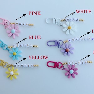 May include: A set of 8 personalized flower keychains with different colored flowers and names spelled out in gold beads. The colors include pink, white, lilac, blue, yellow, and dark pink.