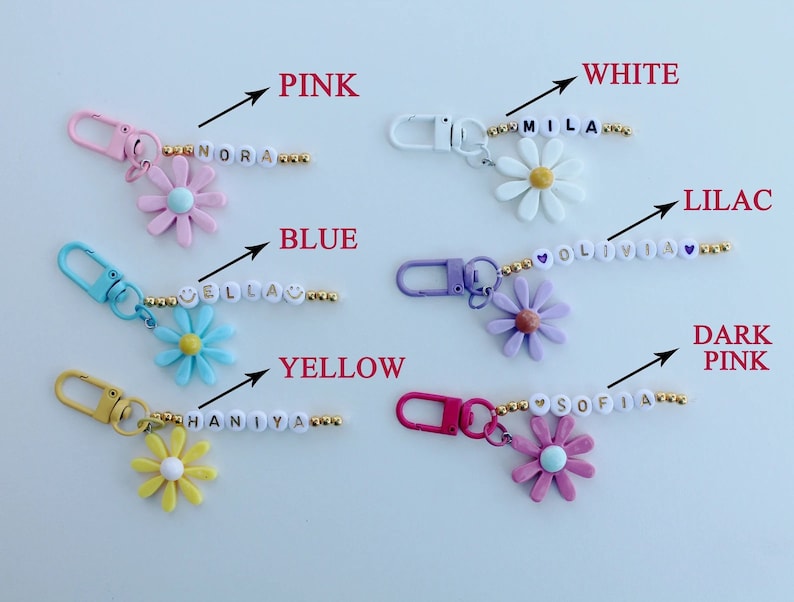 May include: A set of 8 personalized flower keychains with different colored flowers and names spelled out in gold beads. The colors include pink, white, lilac, blue, yellow, and dark pink.