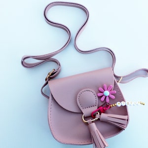 May include: A dusty rose-colored faux leather satchel with a gold clasp and a pink flower charm. The bag has a long adjustable strap and a tassel. The bag has a beaded charm with the word "Sofia" on it.