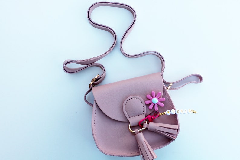 May include: A dusty rose-colored faux leather satchel with a gold clasp and a pink flower charm. The bag has a long adjustable strap and a tassel. The bag has a beaded charm with the word "Sofia" on it.