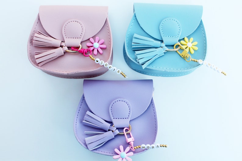 May include: Three small, faux leather purses with a flap closure and a tassel. Each purse has a gold-colored chain with a flower charm and a beaded name tag. The purses are pink, blue, and purple.
