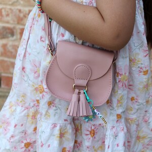May include: A pink faux leather crossbody bag with a gold clasp and tassel charm. The bag is worn by a child wearing a white floral dress.