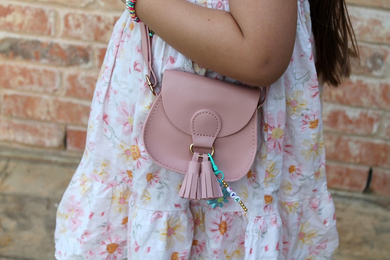 May include: A pink faux leather crossbody bag with a gold clasp and tassel charm. The bag is worn by a child wearing a white floral dress.