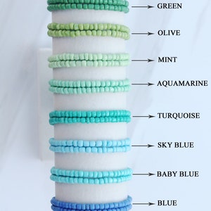 Op de afbeelding: Een set van tien kralenarmbanden in verschillende tinten groen, blauw en zwart. De armbanden zijn verticaal gerangschikt, met de donkerste kleur onderaan en de lichtste kleur bovenaan. De kleuren zijn van boven naar beneden gelabeld: Forest, Green, Olive, Mint, Aquamarine, Turquoise, Sky Blue, Baby Blue, Blue en Black.