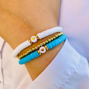 Pode incluir: Tr&ecirc;s pulseiras empilhadas em um pulso. A pulseira superior &eacute; branca com pingentes de cora&ccedil;&atilde;o dourados. A pulseira do meio &eacute; de contas douradas. A pulseira inferior &eacute; azul turquesa com pingentes de cora&ccedil;&atilde;o dourados.