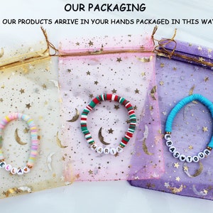Op de afbeelding: Drie kleurrijke kralenarmbanden met gepersonaliseerde namen, Ella, Maria en Erastour, verpakt in individuele mesh-zakjes met een sterpatroon.