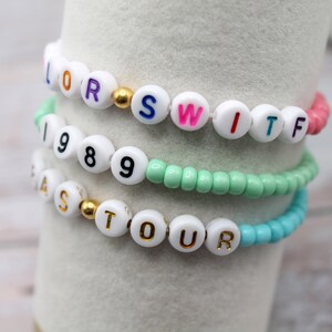 Op de afbeelding: Vier kralenarmbanden met witte kralen en kleurrijke letters die "LORS", "SWITF", "1989" en "FASTOUR" spellen.