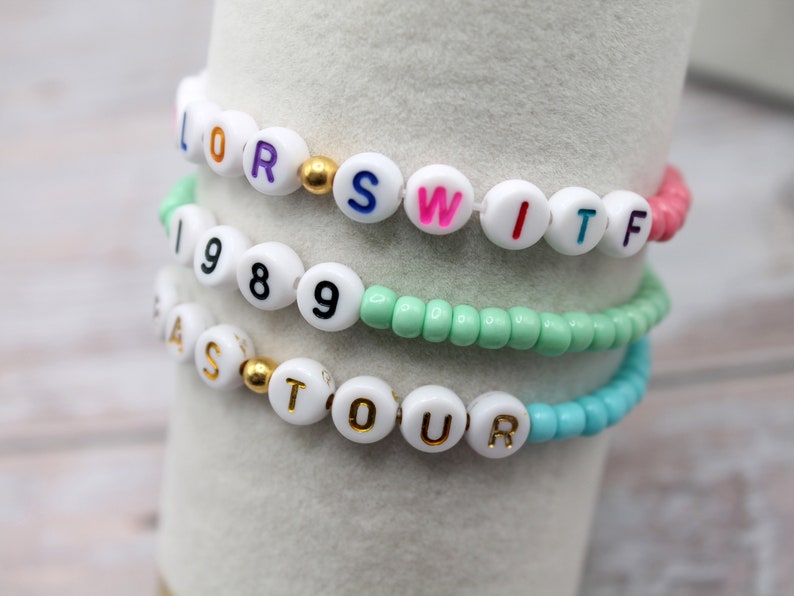 Op de afbeelding: Vier kralenarmbanden met witte kralen en kleurrijke letters die "LORS", "SWITF", "1989" en "FASTOUR" spellen.
