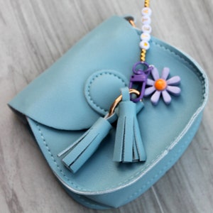 May include: A small blue faux leather purse with a flower charm and tassels. The purse has a gold chain strap and a white beaded charm.