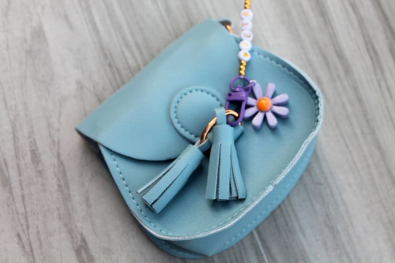 May include: A small blue faux leather purse with a flower charm and tassels. The purse has a gold chain strap and a white beaded charm.
