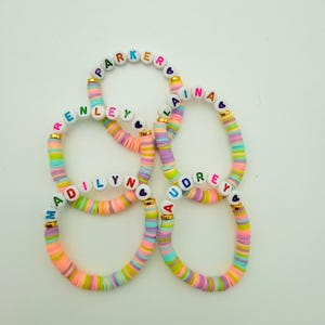 Pode incluir: Cinco pulseiras coloridas com contas brancas e letras coloridas que soletram os nomes "Parker", "Alana", "Renley", "Madilyn" e "Audrey". Cada pulseira tem um pequeno berloque de cora&ccedil;&atilde;o dourado.