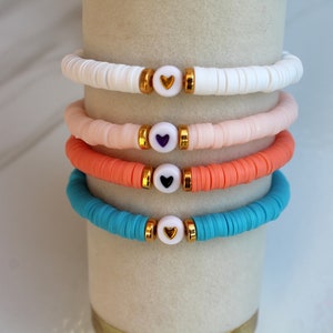 May include: Four colorful bracelets with gold heart charms. The bracelets are stacked on top of each other. The top bracelet is white, the second is pink, the third is orange, and the bottom bracelet is turquoise.