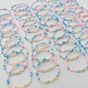 May include: A collection of elastic bracelets made from small round beads in alternating light blue, pink, and white colors. Each bracelet features white letter beads spelling DSM with a small gold heart symbol on either side of the letters.
