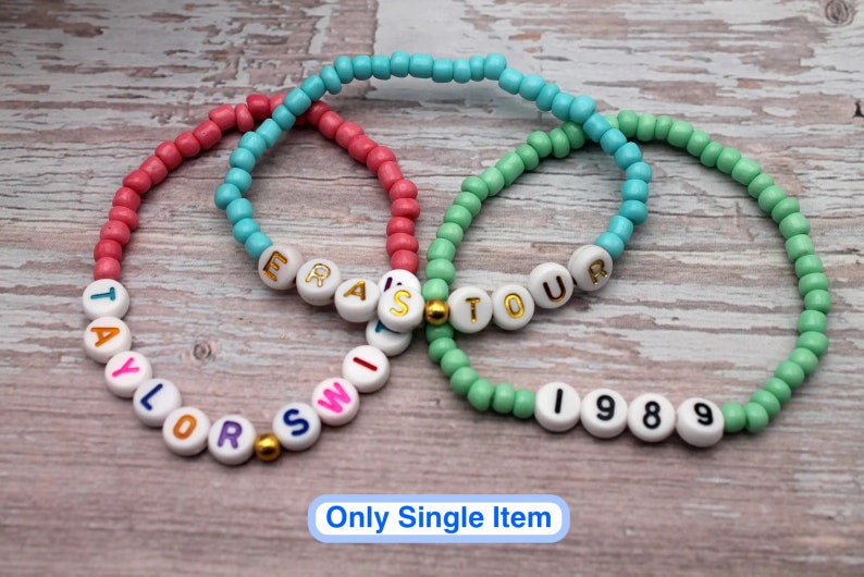 Op de afbeelding: Drie kralenarmbanden met witte kralen en gouden letters. De armbanden spellen "Taylor Swift", "Eras Tour" en "1989".