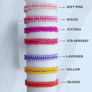 Op de afbeelding: Een set van tien kralenarmbanden in verschillende kleuren, waaronder wit, zacht roze, rood, fuchsia, aardbei, lavendel, geel, oranje en rood. De armbanden zijn verticaal op een witte achtergrond gerangschikt, met de kleurennamen naast elke armband.